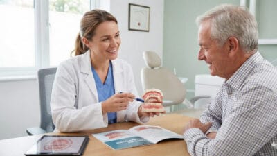 Female dentist showing a complete dentures model to a senior male patient during a consultation, explaining denture types, fitting, and daily care.